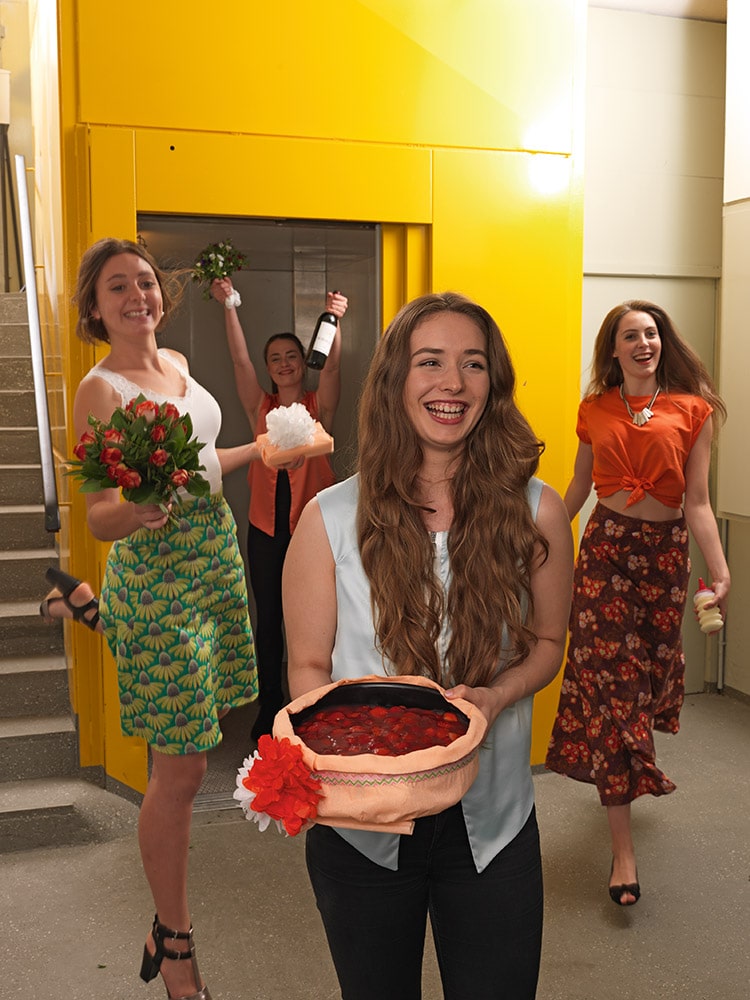 Vier Frauen bei einer Feier, eine hält einen Blumenstrauß, eine andere einen Kuchen, alle lachen und tragen bunte, kreative Kleidung, im Hintergrund eine gelbe Wand.